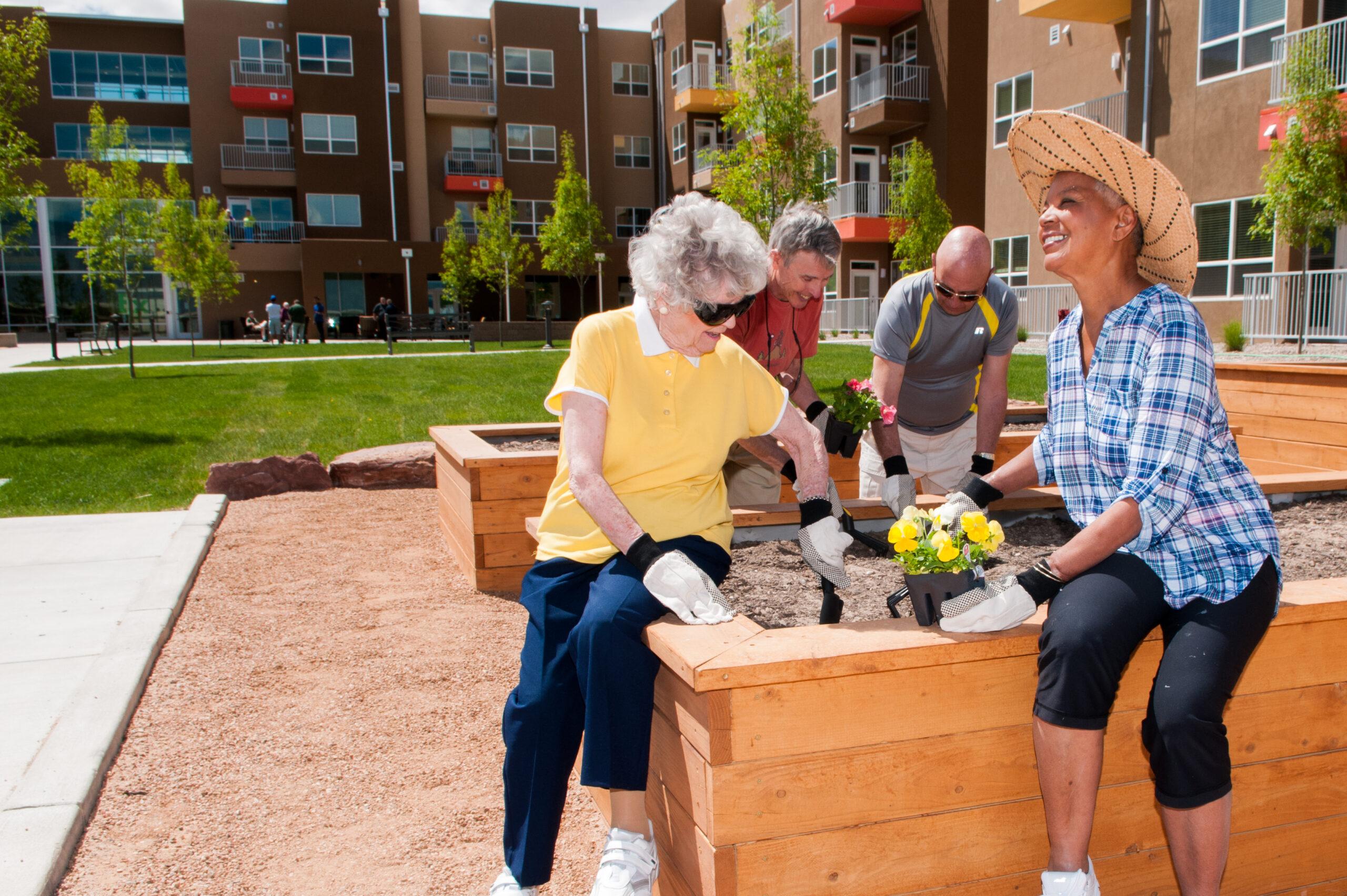 A diverse group of seniors planting flowers in raised garden beds outside an apartment complex on a sunny day.
