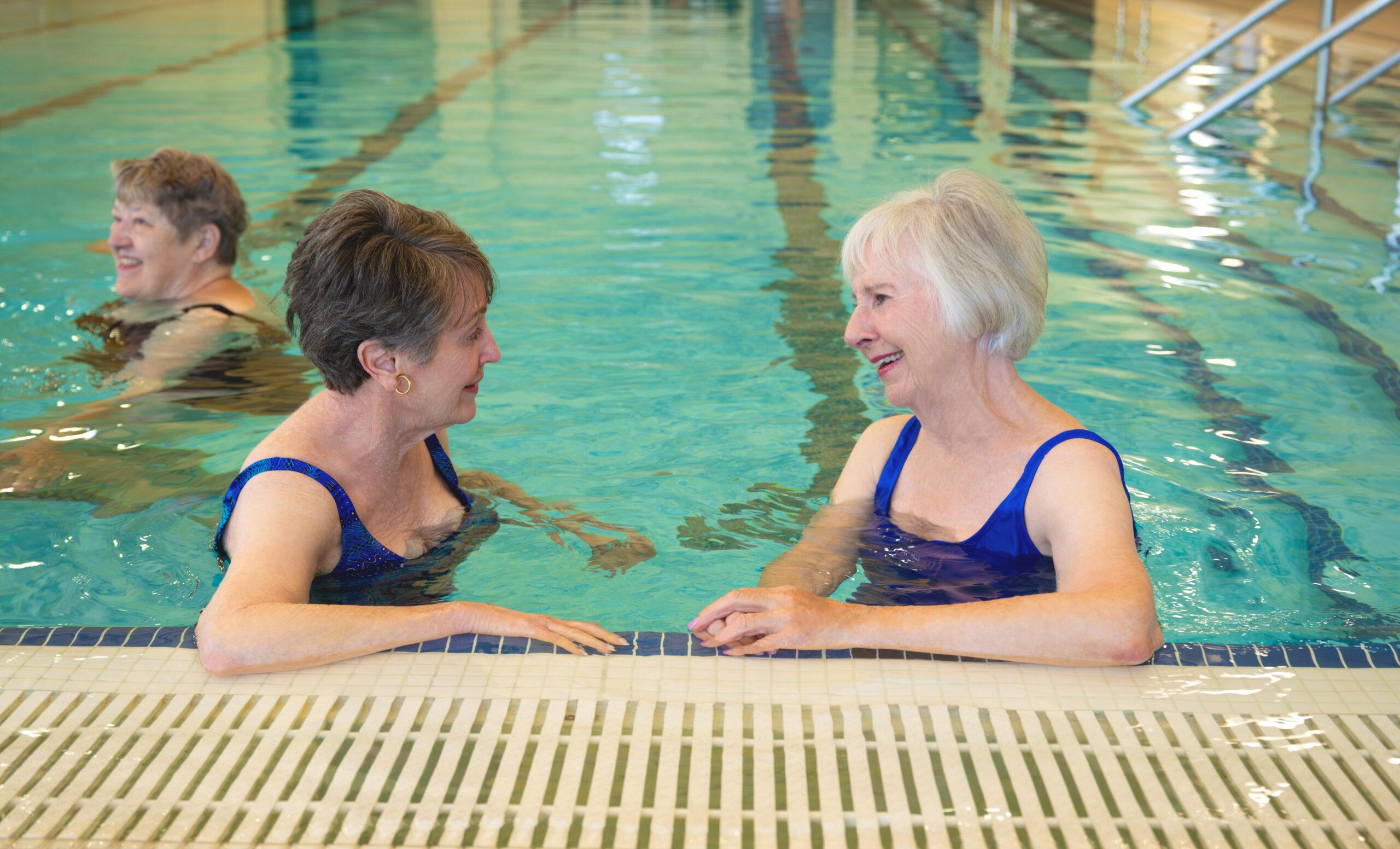 Two senior women conversing while standing waist-deep in a swimming pool, with other swimmers behind them.