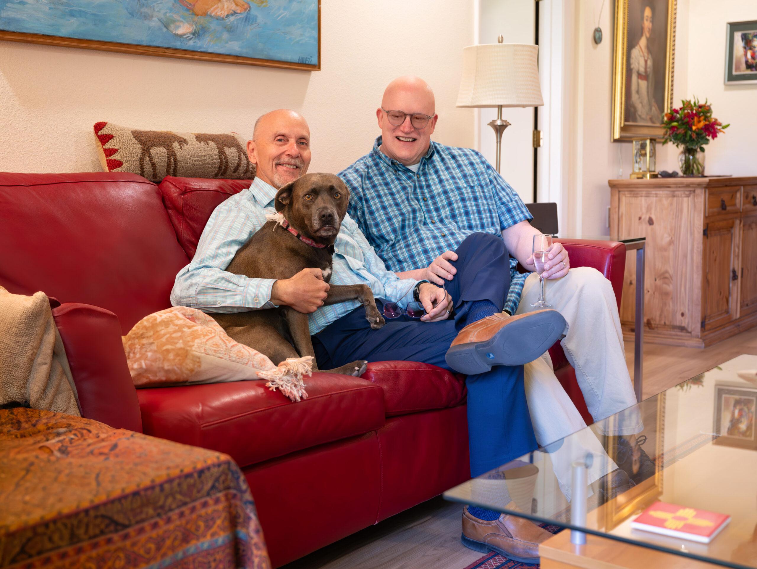 Two middle-aged men sitting on a red leather sofa, one holding a brown dog, both smiling and holding wine glasses in a living room.