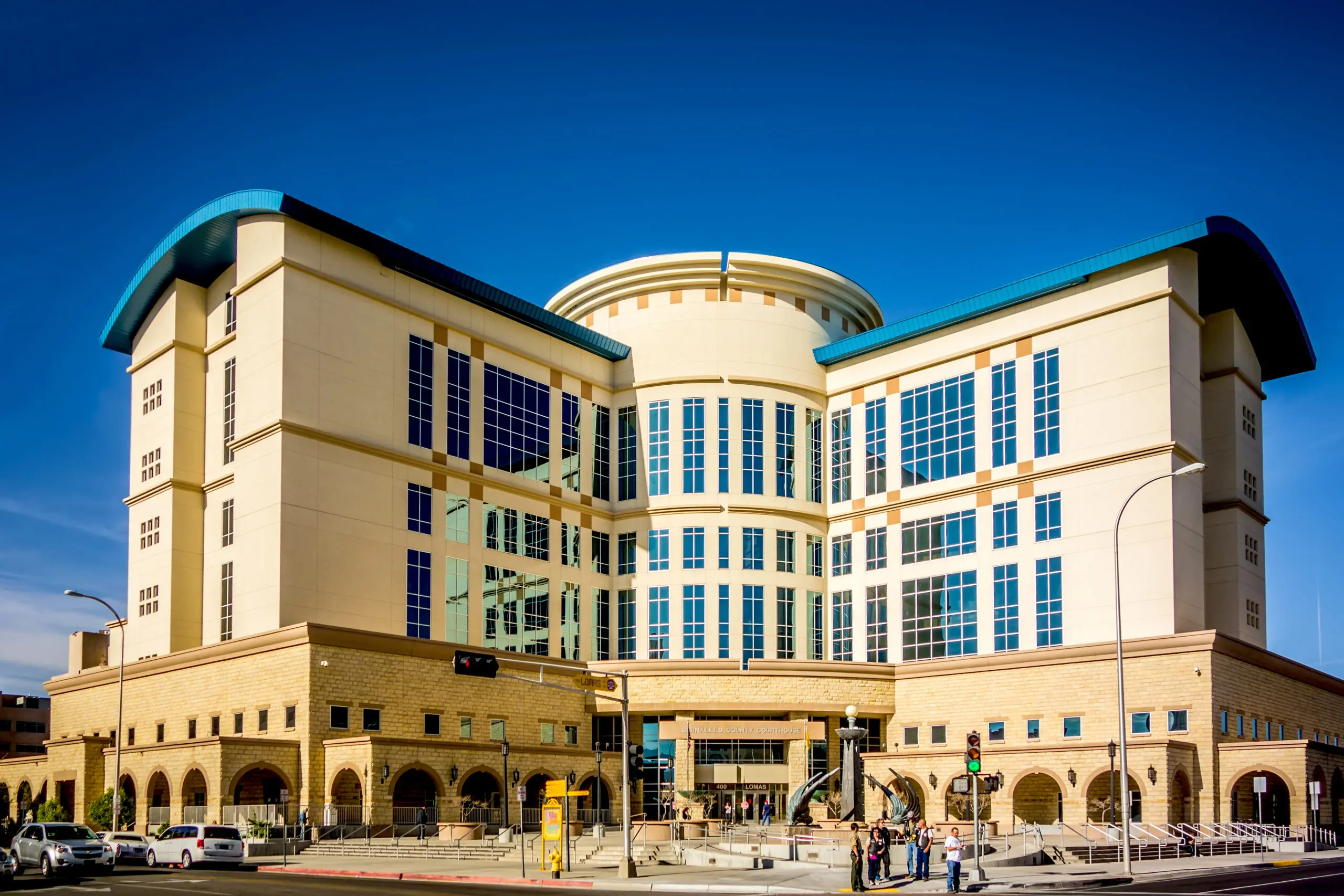 The beige and turquoise-roofed Bernalillo County Courthouse, a modern stucco building with arched ground-floor openings, on a sunny city street corner.