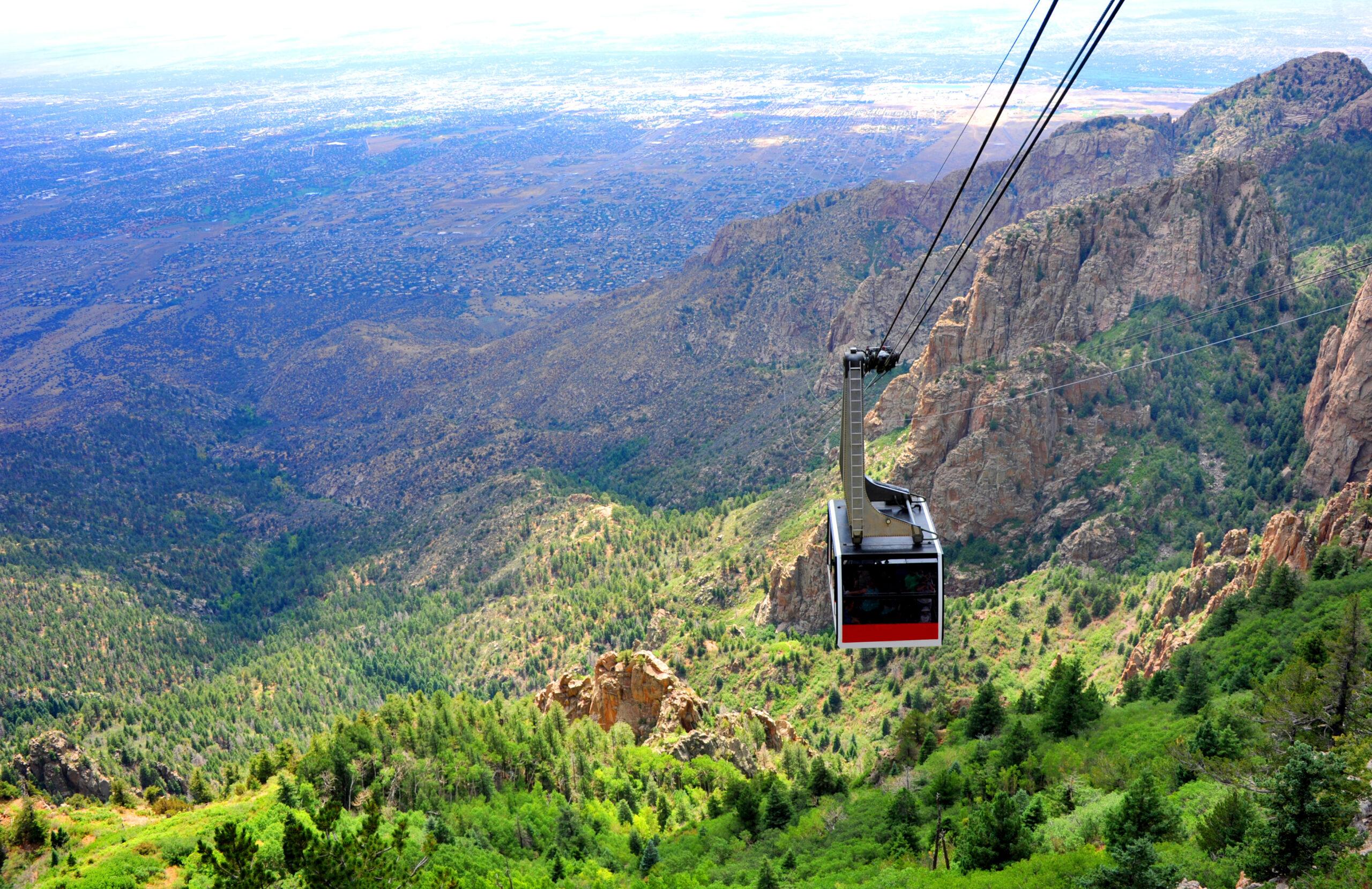 A red Sandia Peak Tram car suspended by cables over a forested mountainside, with the sprawling Albuquerque valley visible far below.